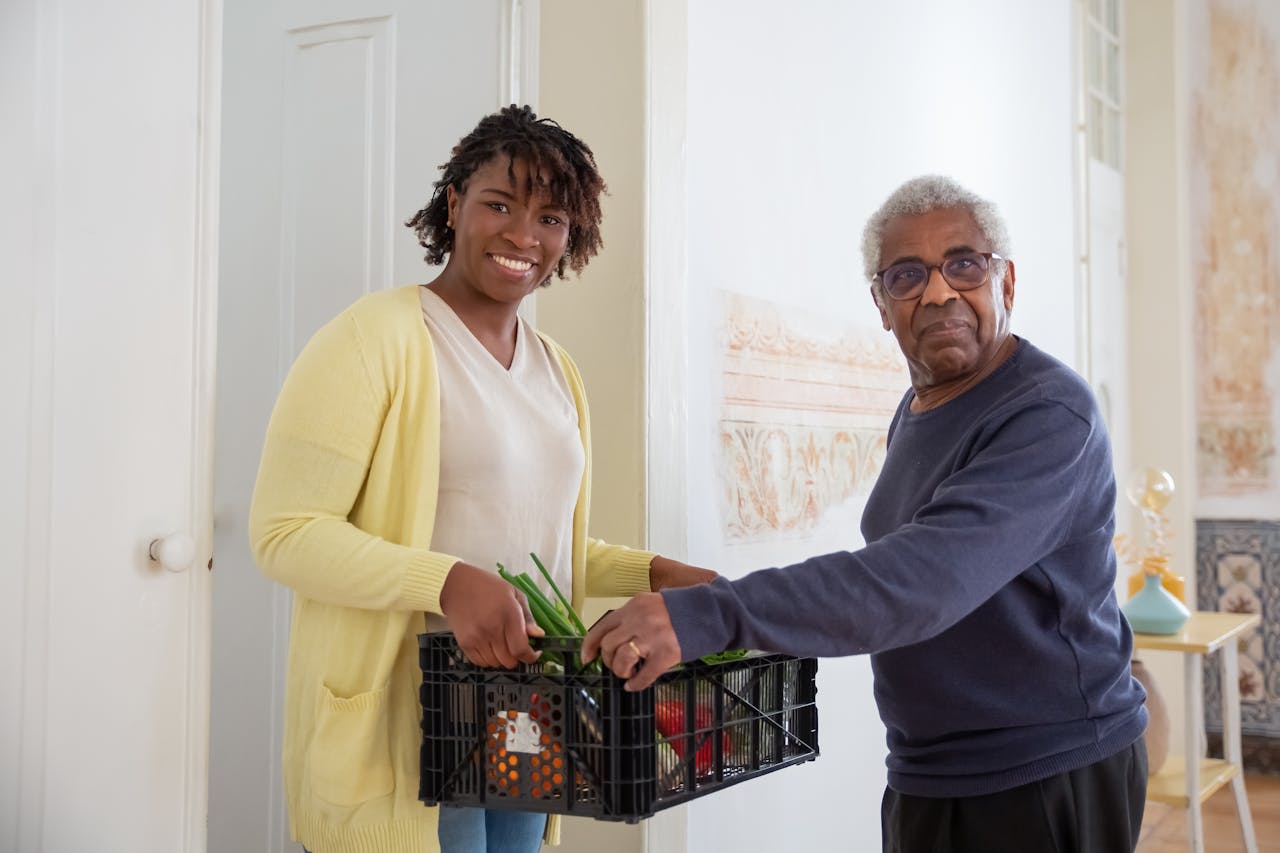 Smiling woman assists a senior man with food delivery indoors, promoting community support and care.